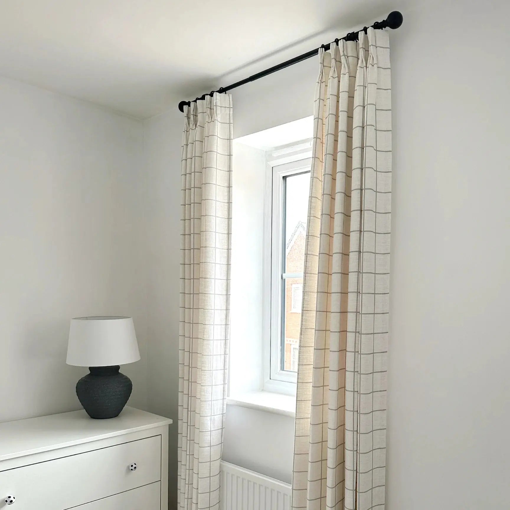 A bright bedroom interior featuring a window with beige grid-patterned curtains on a black rod, a white dresser with soccer ball knobs, and a dark grey textured lamp with a white shade