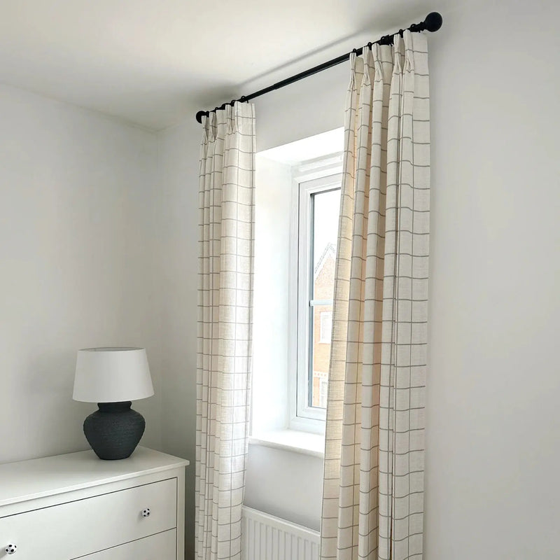 A bright bedroom interior featuring a window with beige grid-patterned curtains on a black rod, a white dresser with soccer ball knobs, and a dark grey textured lamp with a white shade