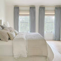 Clean modern bedroom showcasing light gray pleated drapes on brass rods, a bed dressed in crisp white bedding with elegant lace trim, and an array of textured neutral pillows before three large sunlit windows
