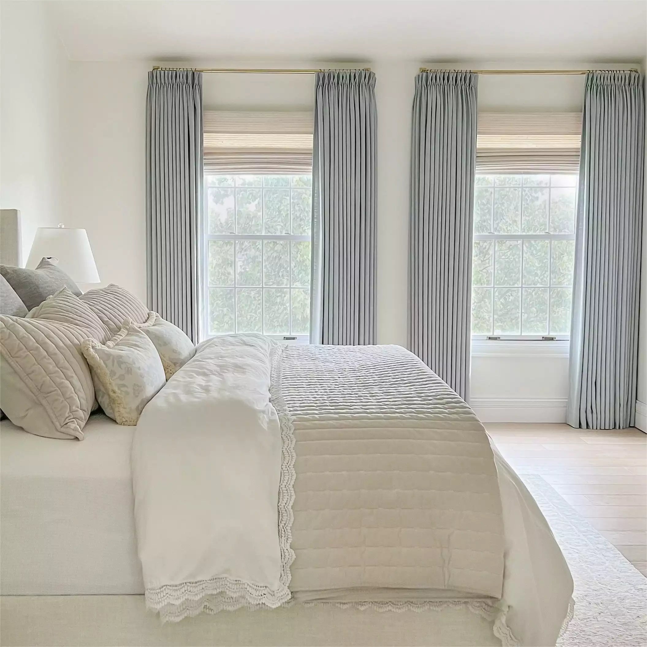 Clean modern bedroom showcasing light gray pleated drapes on brass rods, a bed dressed in crisp white bedding with elegant lace trim, and an array of textured neutral pillows before three large sunlit windows