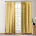A pair of golden yellow pleated linen-blend curtains hangs from a black rod in a bright room with a window overlooking green trees, beside a ribbed wooden side table holding a black vase with greenery and a gold candlestick