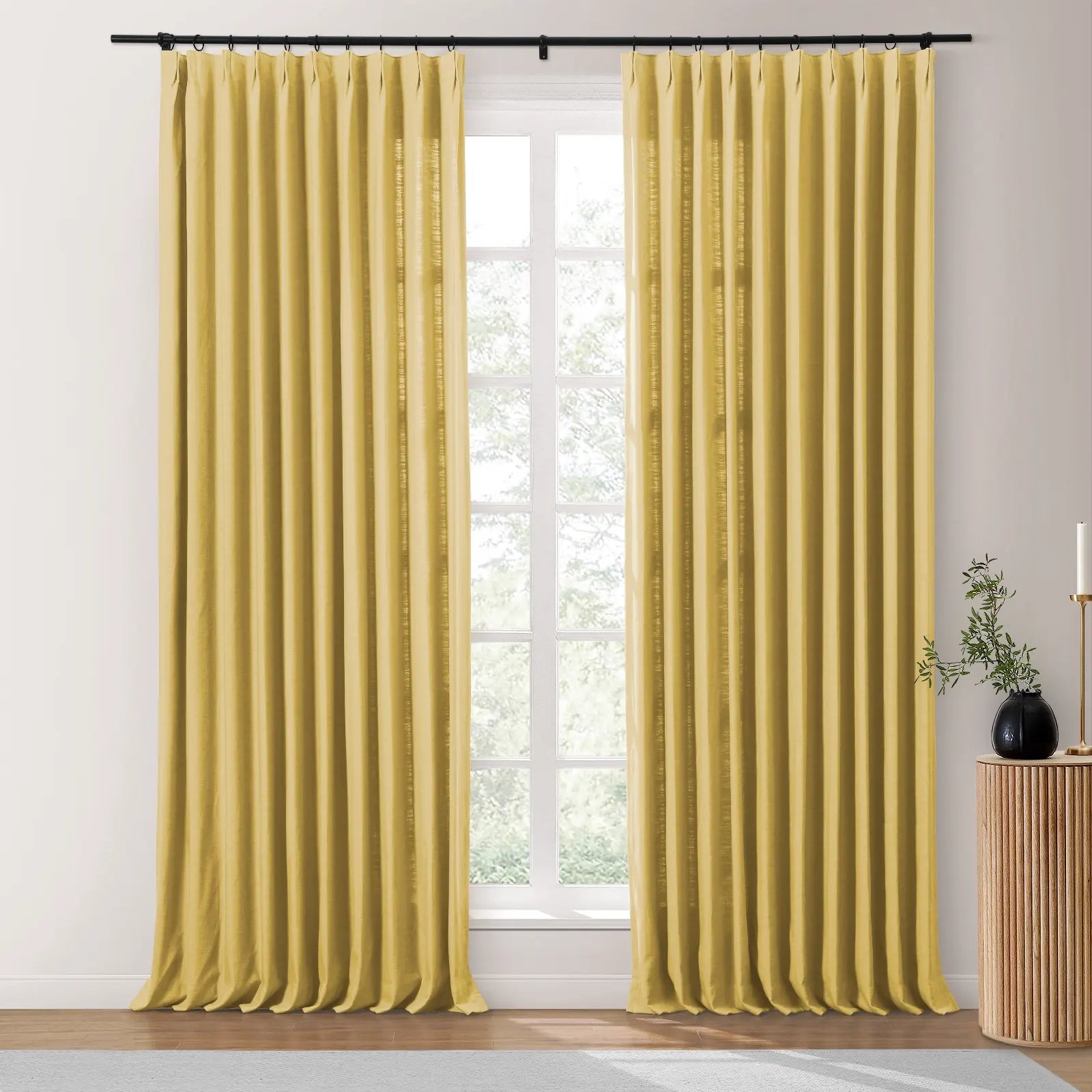 A pair of golden yellow pleated linen-blend curtains hangs from a black rod in a bright room with a window overlooking green trees, beside a ribbed wooden side table holding a black vase with greenery and a gold candlestick