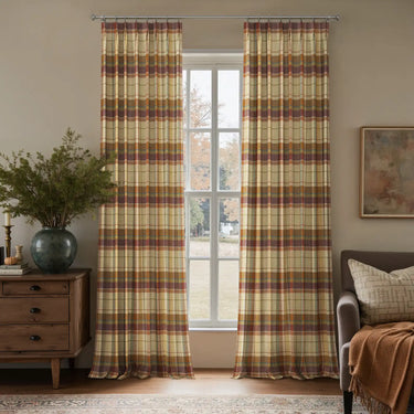 A cozy room interior featuring a large window flanked by full-length plaid curtains in earthy yellows, browns, maroons, and oranges, a rustic wooden dresser adorned with a teal vase holding green foliage, and a grey armchair draped with an orange knit throw blanket overlooking an autumn landscape